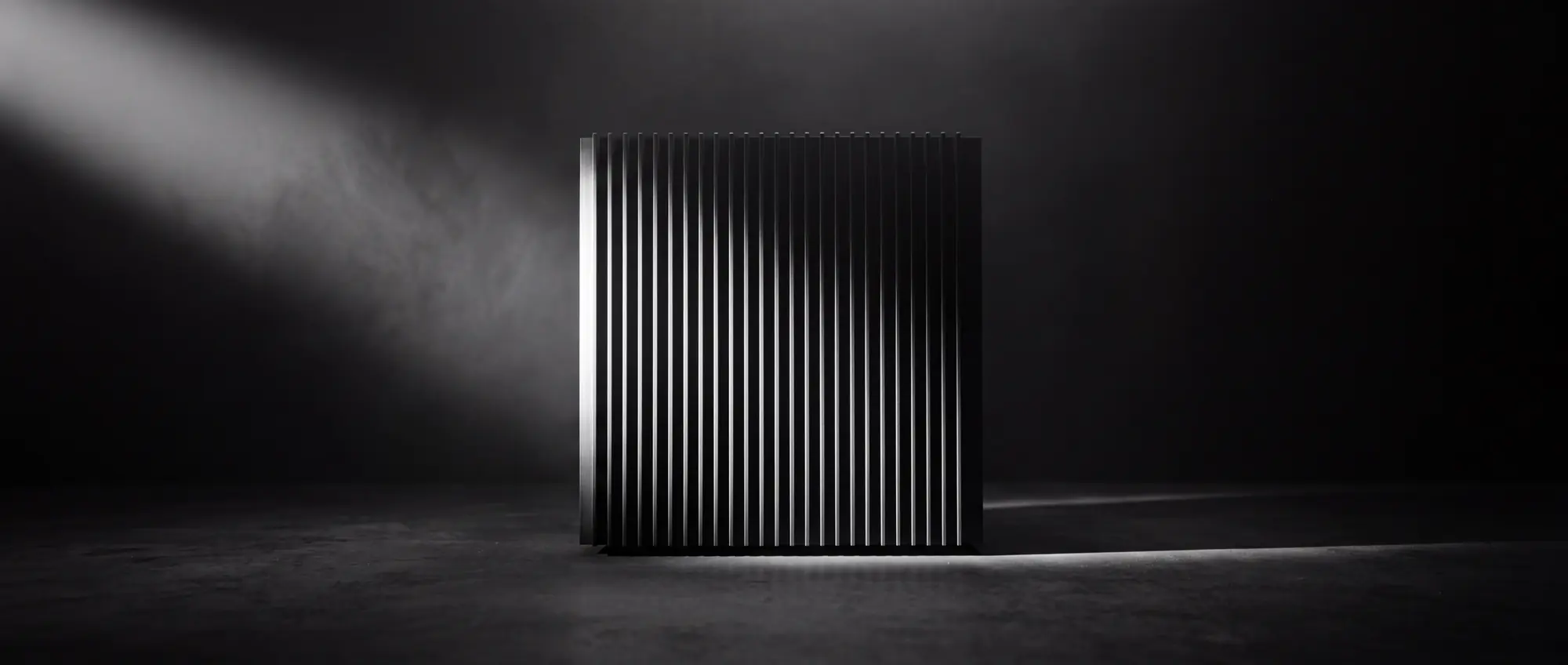 A monolithic finned black compute structure standing in a dark chamber, cut by a single blade of hard light