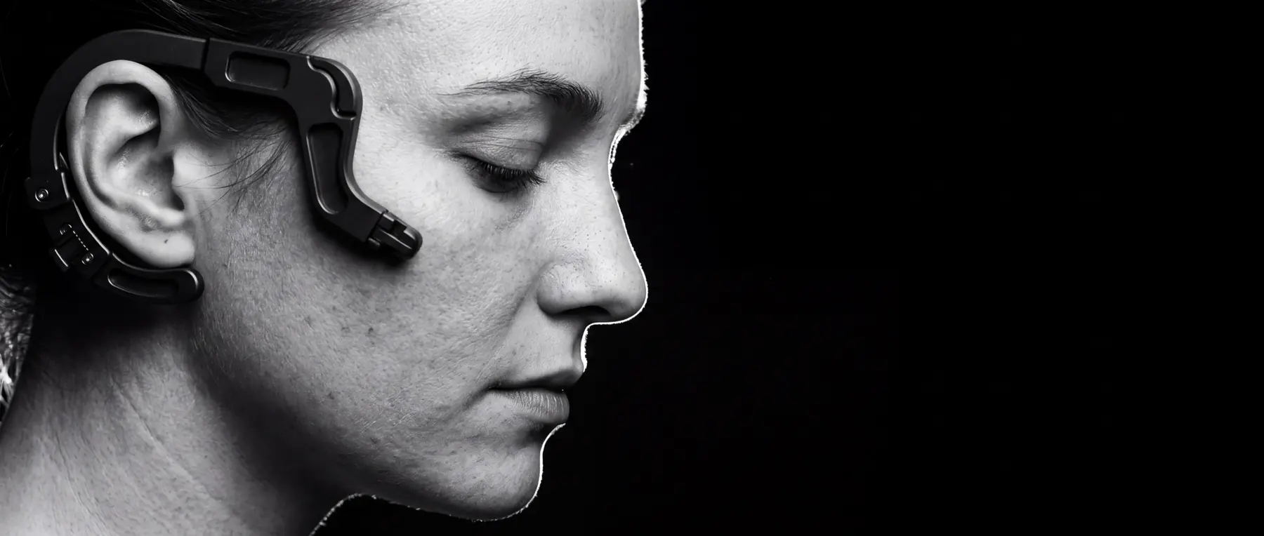 Close profile of a person in near darkness, a matte black machined apparatus curving along the temple